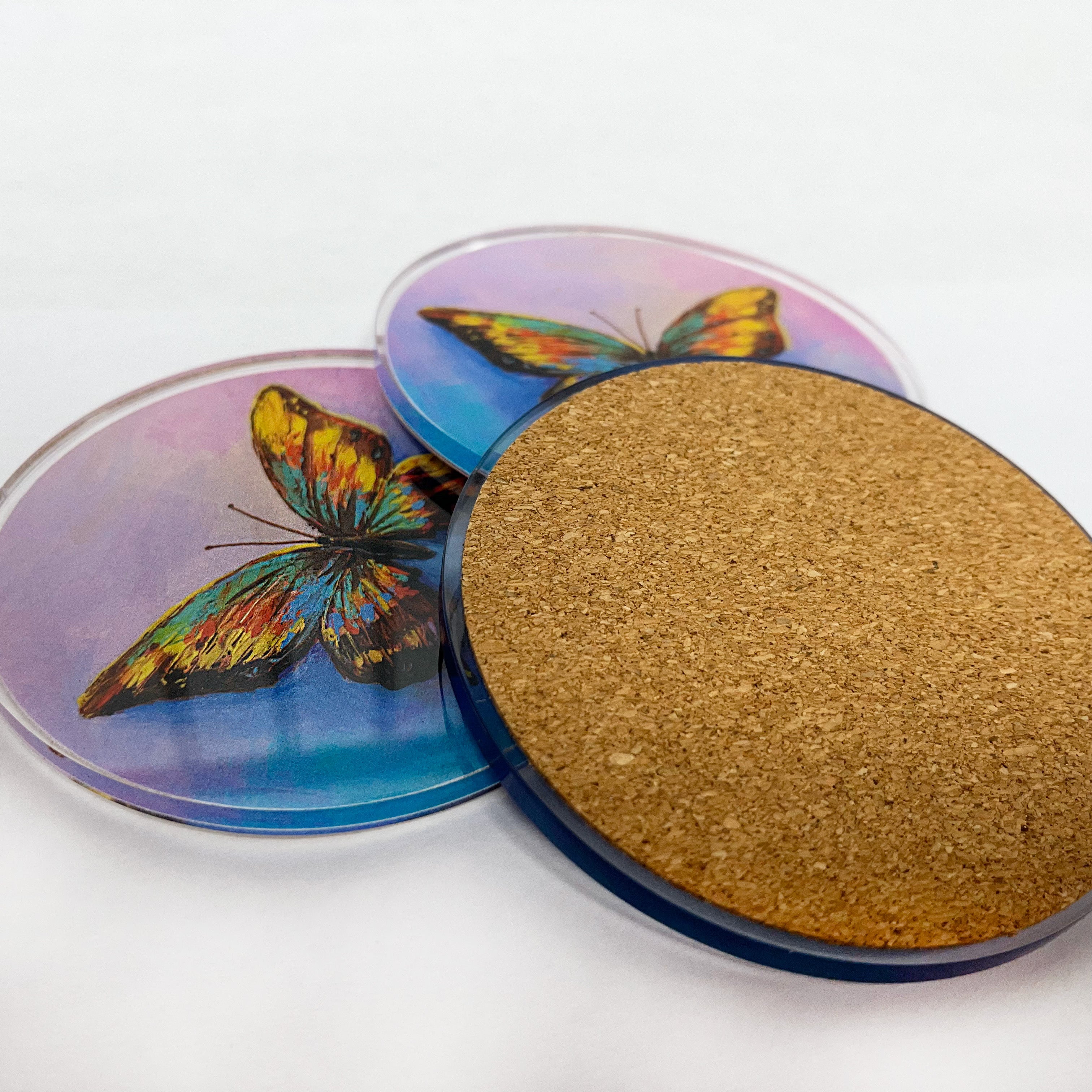 3 acrylic coasters of printed artwork laying on a table with one turned over to show cork backer.